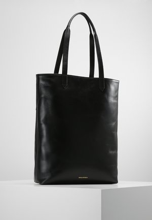 Zwarte leren totebag met twee schouderbanden, rechtopstaand op een wit voetstuk tegen een effen achtergrond.