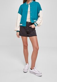 Młoda kobieta w turkusowo-białej bomberce, beżowej bluzce, czarnych szortach oraz białych trampkach na platformie, stoi z jedną ręką w kieszeni.