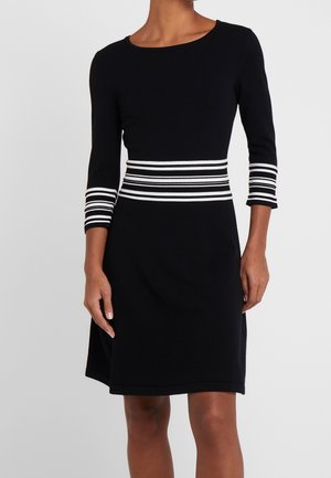 Černé šaty délky po kolena s tříčtvrtečními rukávy a bílými vodorovnými pruhy na pasu a manžetách, nošené stojící osobou.