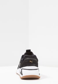 Chaussure de sport noire avec une tige en mesh texturé, une semelle intermédiaire blanche, une semelle extérieure en caoutchouc marron et un logo doré détaillé sur le talon.