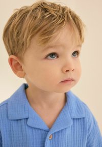 Camisa azul clara, texturizada, con cuello y detalles de botones de madera. Niño con cabello rubio corto y despeinado y ojos azules.