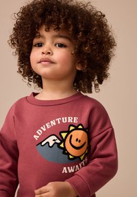 Vyno raudonos spalvos džemperis su ryškiu saulės piešiniu, kurioje matyti besišypsanti veidas, kalnas ir baltomis raidėmis užrašytas tekstas „ADVENTURE AWAITS“. Minkšta medžiaga.