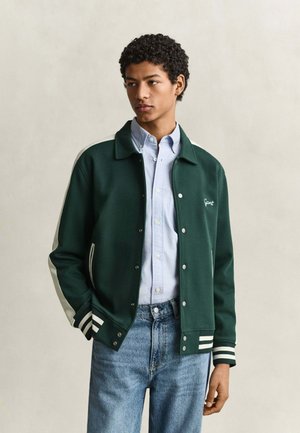 Junger Mann trägt eine dunkelgrüne Collegejacke über einem hellblauen Hemd und blauen Jeans, steht mit der Hand in der Tasche und schaut seitlich.