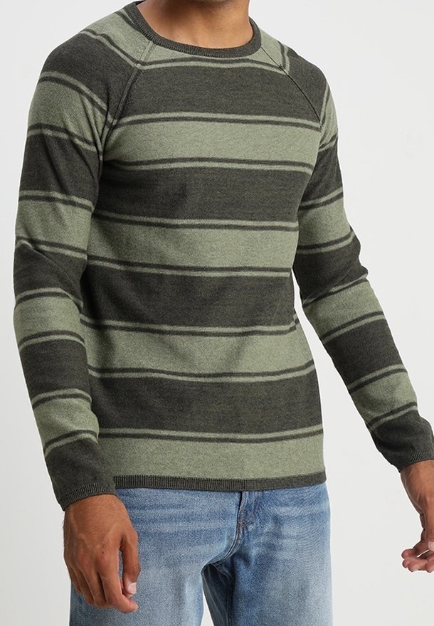 Homme portant un pull à manches longues rayé vert et gris foncé avec un jean bleu clair, debout devant un fond uni.