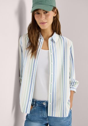 Kobieta w zielonej czapce, białym topie na ramiączkach, luźnej, paskowanej koszuli zapinanej na guziki i niebieskich dżinsach, stojąca z rękami w kieszeniach na tle jasnej ściany.