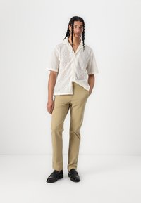 Chemise blanche à manches courtes avec un motif en zigzag, associée à un pantalon beige et des chaussures noires, se tenant devant un fond blanc.