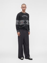 Homme portant un pull noir et gris avec un motif de lapin, un pantalon ample foncé et des chaussures noires et blanches, debout devant un fond uni.