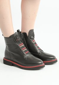 Bottines en cuir noir avec des accents rouges, fermeture éclair sur le côté et panneaux élastiques rayés avec des motifs en chevrons rouges et roses. Semelle durable.