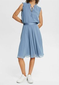 Vaaleansininen mekko, jossa on hihaton yläosa, jossa on röyhelöreunat, säädettävät nauhat kaula-aukossa ja virtaava A-linjainen hame.