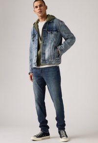 Denim jacket in light blue, layered over an olive green hooded sweatshirt, paired with dark blue jeans and black low-top sneakers.