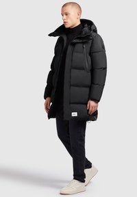 Svart vadderad vinterjacka med huva, dragkedja framtill och sidofickor, med en strukturerad yta och logotypdetalj nära kanten.