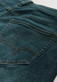 Tumesinistes toonides teksapüksid, millel on tekstuuriga viimistlus. Tagataskul kontrastset kollast õmblust ja peened tuhmid detailid.