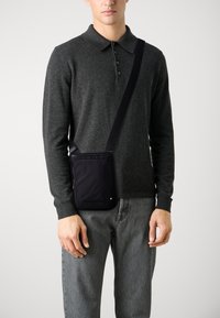 Sort crossbody taske lavet af stof, med en lynlåslomme foran, justerbar rem og minimalistisk design. Båret over en grå sweater.