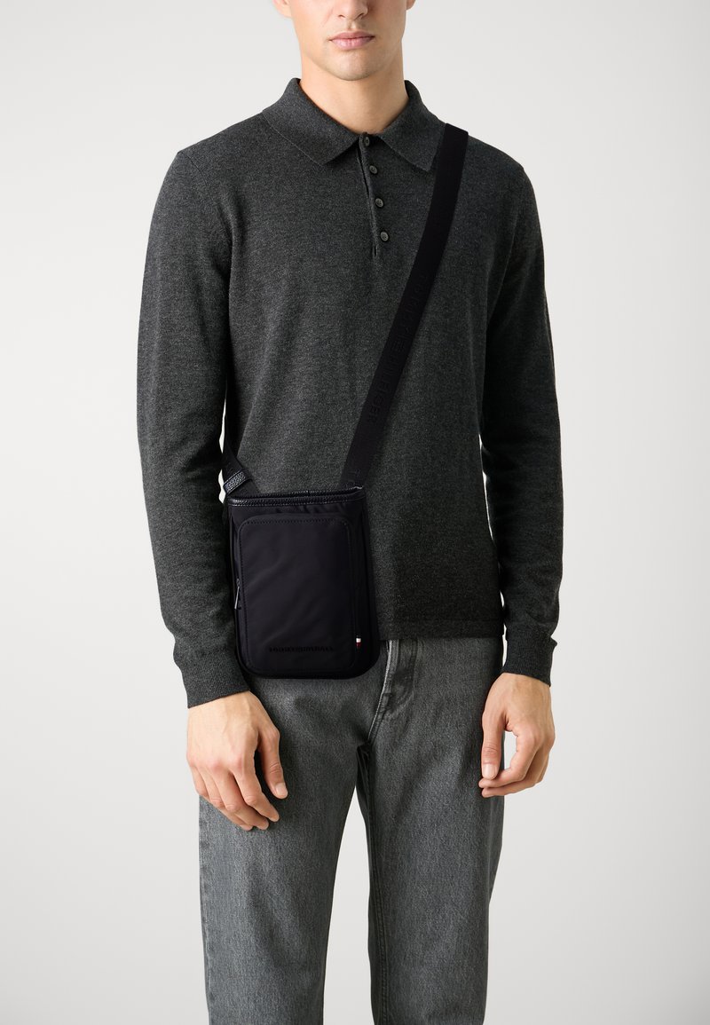 Sort crossbody taske lavet af stof, med en lynlåslomme foran, justerbar rem og minimalistisk design. Båret over en grå sweater.