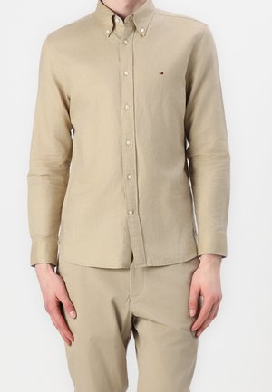 Camicia beige a maniche lunghe con colletto a punta, caratterizzata da un piccolo logo sul petto e un tessuto liscio e texture.