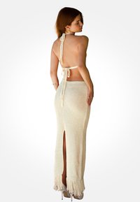 Mujer con cabello rojo que lleva un vestido halter de punto color crema sin espalda con borde de flecos y tacones altos, mirando por encima del hombro.