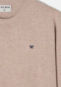 Suéter de punto beige con cuello redondo, puños acanalados y un pequeño emblema de tijeras en azul marino en el pecho. Textura suave y diseño sencillo.