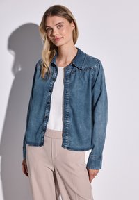 Camisa de mezclilla en azul claro, con cierre de botones en el frente, cuello puntiagudo y mangas largas con puños de botón, superpuesta a una blusa blanca.