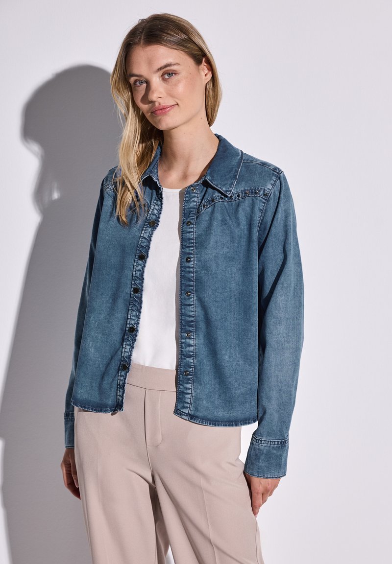 Camisa de mezclilla en azul claro, con cierre de botones en el frente, cuello puntiagudo y mangas largas con puños de botón, superpuesta a una blusa blanca.