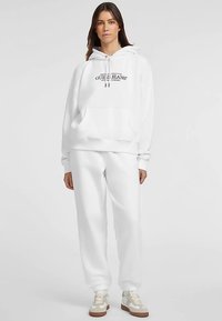 Valge kapuutsiga pusa rinnal logoga, kombineeritud sobivate valgete dressipükstega. Materjal tundub pehme ja lõdvestunud istuvusega.