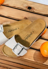 White textured sandals with a silver buckle, featuring a brown suede insole, placed on wood alongside an orange.