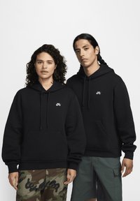 Fekete kapucnis pulóverek elülső zsebekkel, fehér Nike logókkal. A modellek camouflált és cargo sortokat viselnek, kiemelve a különböző stílusokat.