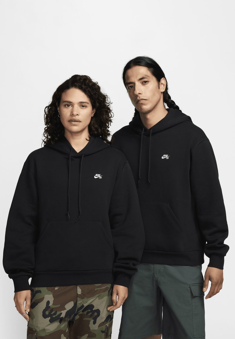 Fekete kapucnis pulóverek elülső zsebekkel, fehér Nike logókkal. A modellek camouflált és cargo sortokat viselnek, kiemelve a különböző stílusokat.