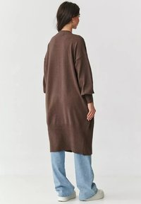 Brązowy, oversize sweter z dzianiny o prążkowanym wykończeniu u dołu i przy mankietach, w zestawieniu z jasnoniebieskimi spodniami typu wide-leg, na tle neutralnym.