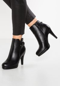 Schwarze Leder-Stiefeletten mit einem hohen Absatz, seitlichem Reißverschluss und einer dekorativen Schnalle. Glatte Textur und elegantes Design.