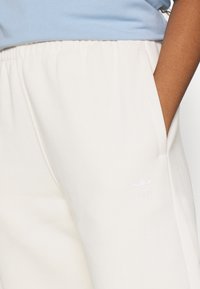 Světle béžové joggingové kalhoty vyrobené z měkké tkaniny, s elastickým pasem, bočními kapsami a bílým vyšívaným logem na levém stehně.