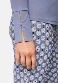 Nahaufnahme einer Hand, die ein blaues Langarmoberteil mit Schlitz und Knopfdetails trägt, kombiniert mit blauen gemusterten Hosen, die weiße und schwarze geometrische Formen zeigen.