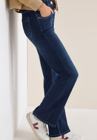 Personne portant un jean en denim bleu foncé et des baskets blanches avec un logo "V" rouge, debout avec une main dans la poche contre un fond beige.