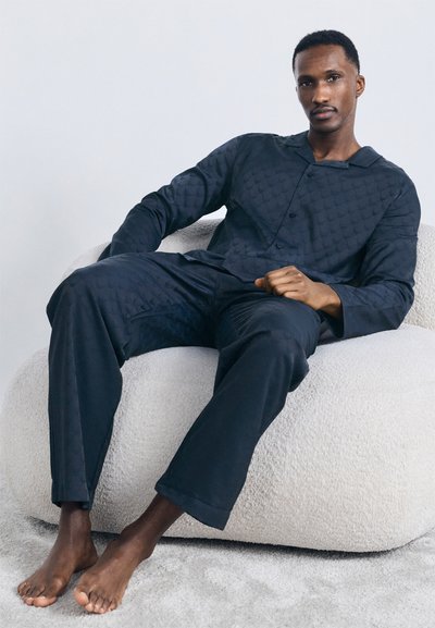Man sitting barefoot on a white textured chair, wearing dark blue patterned long-sleeve top and matching loose pants.