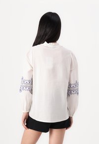 Blusa bianca con maniche a sbuffo, tessuto testurizzato, scollatura arricciata e ricamo floreale blu lungo le maniche. Indossata con pantaloncini neri.