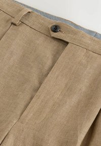 Närbild på midjebandet på beige texturerade byxor med svart knapp och grå fiskbensmönstrad innerfoder.