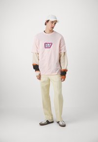 Camiseta rosa con un gran logotipo gráfico estilizado, combinada con pantalones crema y zapatillas. El conjunto resalta las texturas contrastantes de las mangas.