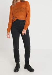 Orange stickad tröja med löst sittande passform och texturerat mönster, kombinerad med svarta figursydda jeans och svarta snörstövlar.