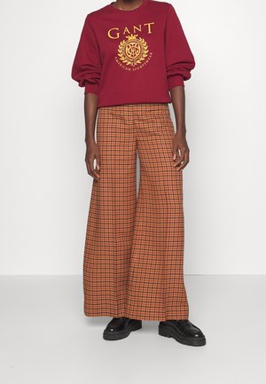 Sudadera burdeos con logo dorado, combinada con pantalones de pata ancha a rayas naranjas y marrones. Unos zapatos de plataforma negros completan el conjunto.