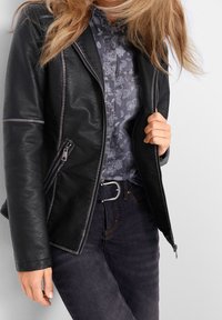 Svart läderbikerjacka med grå sömmar, dragkedjefickor och en något figurnära design, bärs över en mönstrad mörk skjorta och mörka jeans.