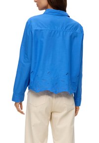 Donna che indossa una blusa a maniche lunghe di un vivace blu con ricami floreali lungo l'orlo, abbinata a pantaloni crema a vita alta, vista da dietro.