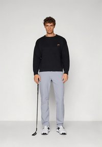 Fekete kötött pulóver kerek nyakkivágással, szürke golfnadrág és sportos cipő. Egy golfütőt tartva a modell semleges háttér előtt áll.