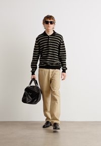 Polo à manches longues noir et olive rayé, pantalon beige coupe ample, lunettes de soleil noires, portant un sac de sport noir, debout sur un sol gris.