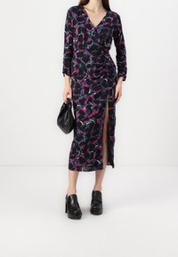 Floral-patterned long dress in navy, pink, and purple, featuring a v-neck, long sleeves, side slit, and soft fabric. Paired with black platform shoes.