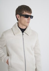 Bežna jakna sa zatvaračem i mekim kragnom od ovčje kože, čiste linije i rebrasti rukavi. Uključena su crna pravokorna sunčana naočala za moderan izgled.