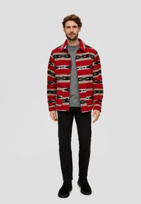 Rot-beige gemusterte Jacke mit schwarzen Akzenten, Knöpfen und einem Kragen, getragen über einem grauen Pullover und einer schwarzen Hose, kombiniert mit schwarzen Schuhen.