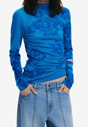 Femme portant un haut bleu vif à manches longues avec des motifs floraux, rentré dans un jean ample en denim bleu clair, les mains dans les poches.