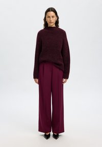 Pull en tricot bordeaux à col montant et manches larges, associé à un pantalon prune ample taille haute et des chaussures noires à bouts pointus.