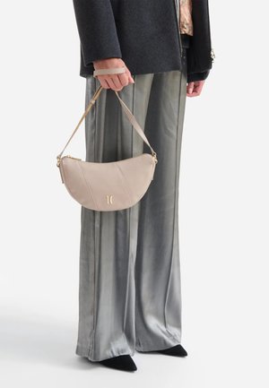 Persona che indossa pantaloni larghi argento, scarpe nere con punta a punta e cappotto scuro, che tiene una piccola borsa beige con logo dorato e tracolla.