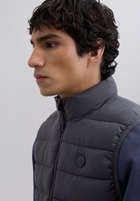 Joven con cabello rizado y oscuro que lleva una chaqueta sin mangas acolchada negra sobre una camisa de manga larga azul oscuro, de perfil izquierdo frente a un fondo liso.