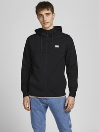 Schwarze Reißverschluss-Hoodie mit verstellbarem Kordelzug für die Kapuze, mit einem Logo auf der Brust und gerippten Bündchen, kombiniert mit hellblauen Jeans.